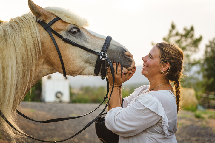 A middle aged woman with blonde hair cups her horses muzzle after a hack