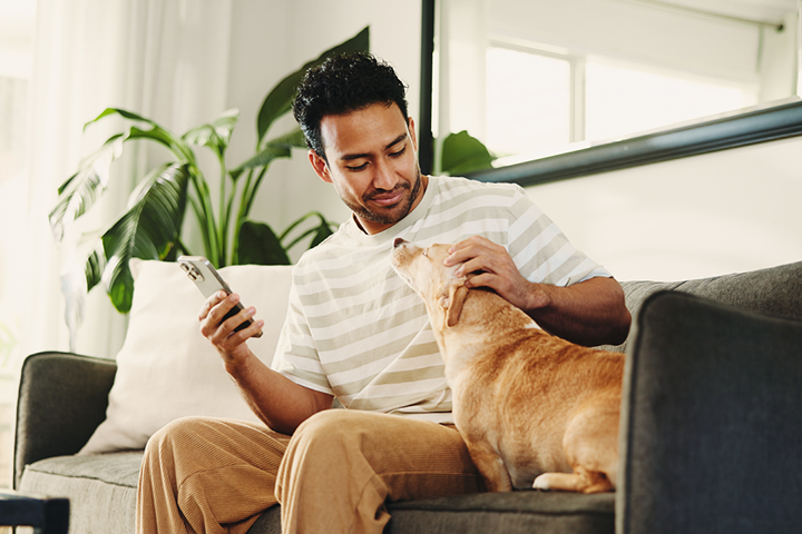 A young mixed-race man sits on the couch with his dog, checking the renewal price for his pet insurance on his phone.